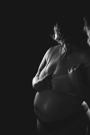 Black and white artistic photo of a pregnant woman cradling her baby bump in dramatic low-key lighting.