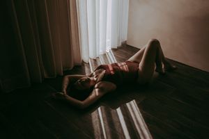 Woman in red lingerie lying on hardwood floor in dramatic light and shadow near sheer curtains.