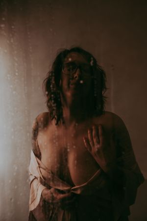 A person with braids and glasses stands behind a wet glass shower door in soft warm moody lighting.