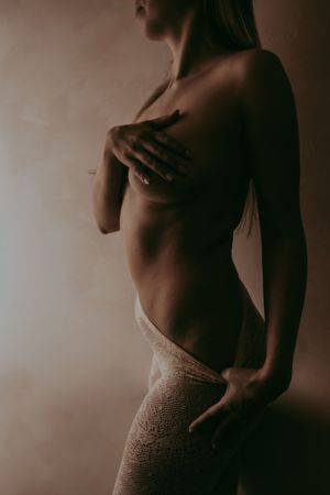 A woman in lace pants poses topless in soft warm lighting, covering her chest with her hand.
