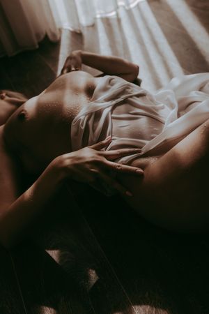 A person lying on a wooden floor draped in sheer white fabric with soft natural light streaming through curtains.