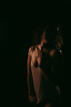 A woman with curly hair poses in a white tank top against a dark background in a dramatic, moody low-light portrait.