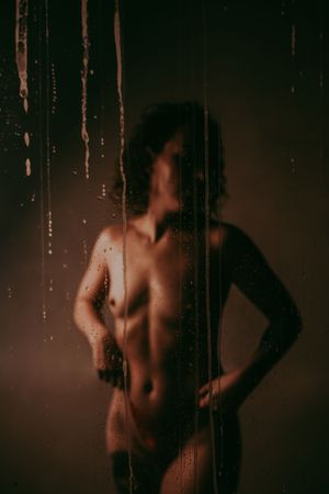 Silhouette of a nude figure behind wet glass with water droplets, captured in warm moody lighting.
