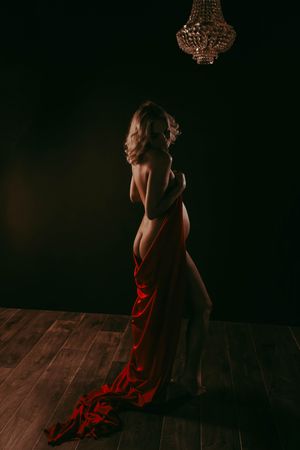 Elegant woman draped in red fabric standing under crystal chandelier in dramatic dark studio lighting.