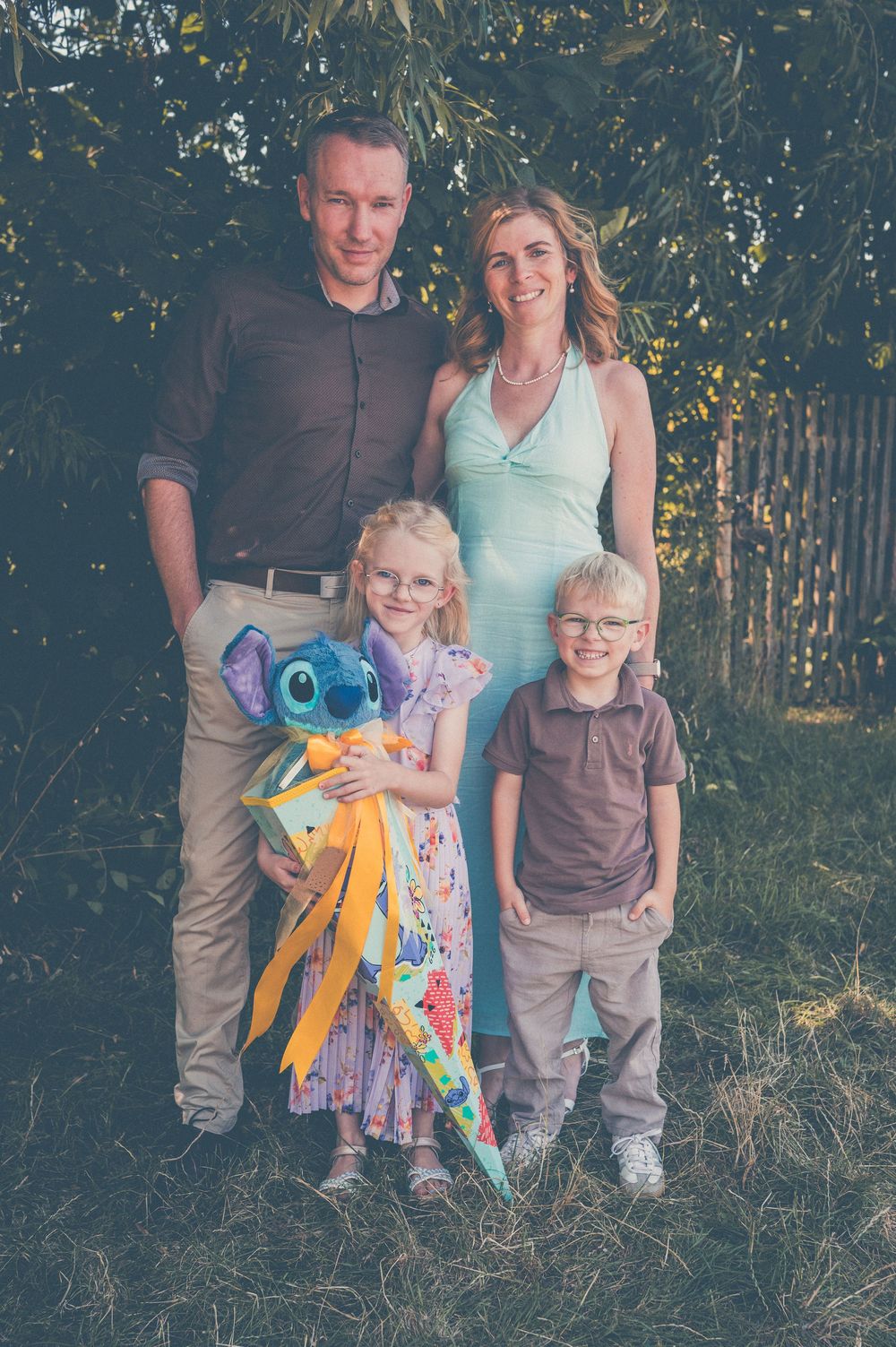 Family of four posing outdoors, girl holding Stitch plush toy, parents and two children smiling in garden setting.