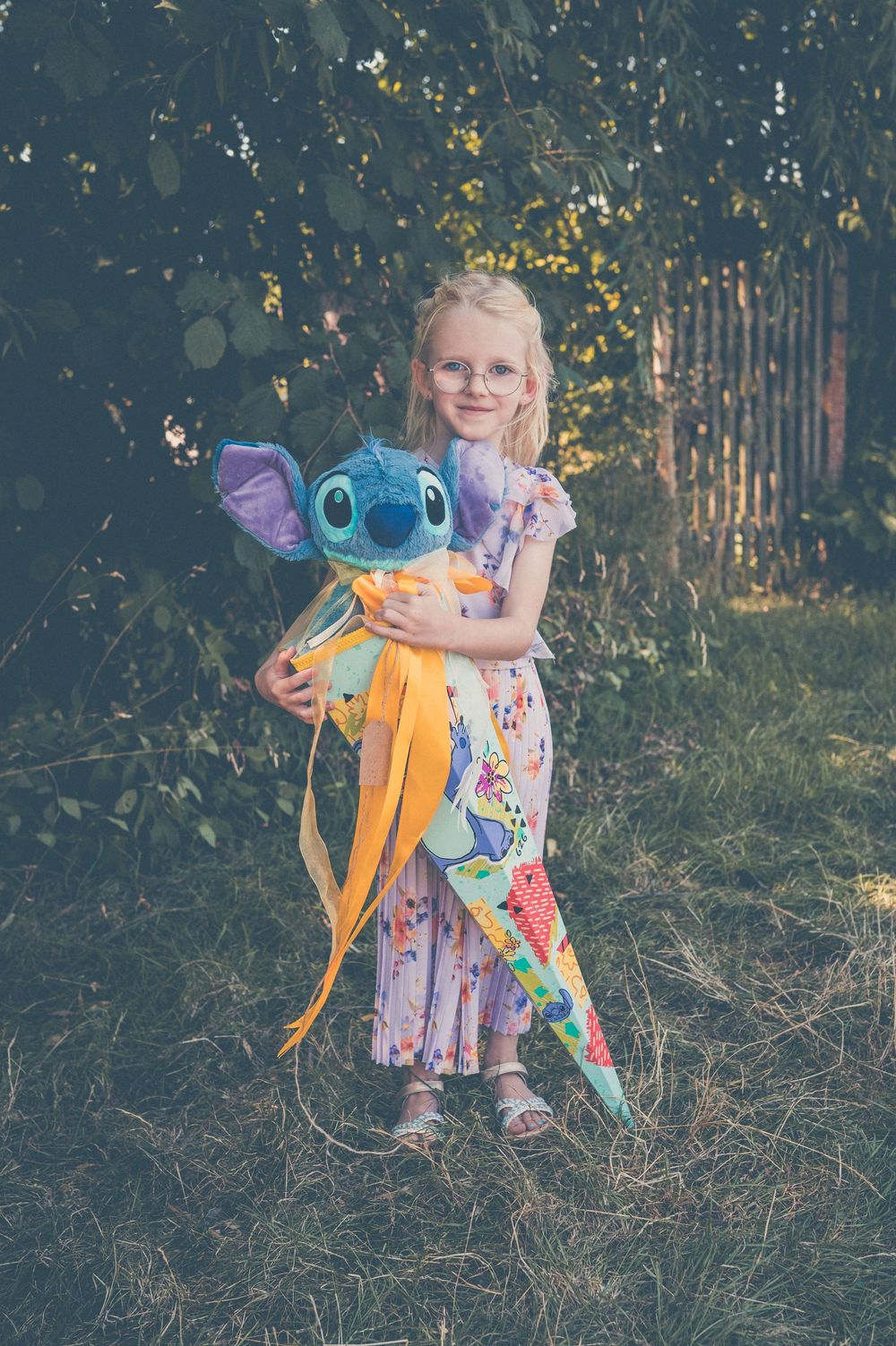 Young blonde girl in floral dress holding a Stitch plush toy and colorful cone gift bag outdoors.