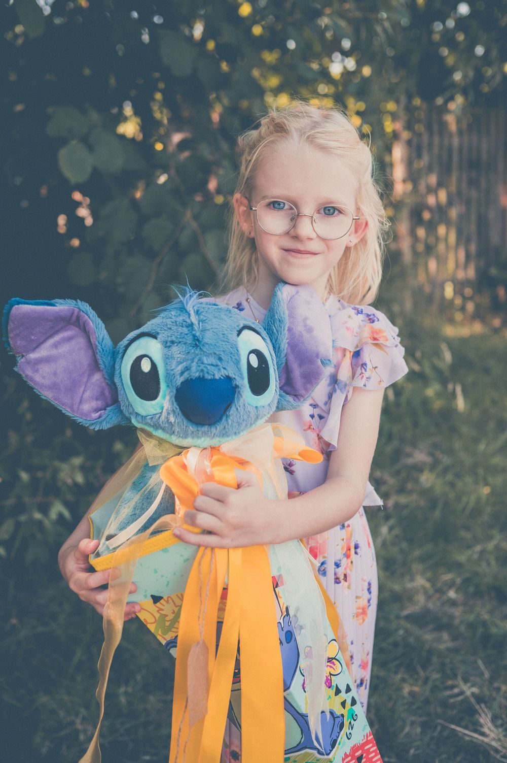 Young blonde girl with glasses hugging a large Stitch plush toy outdoors in a garden setting.