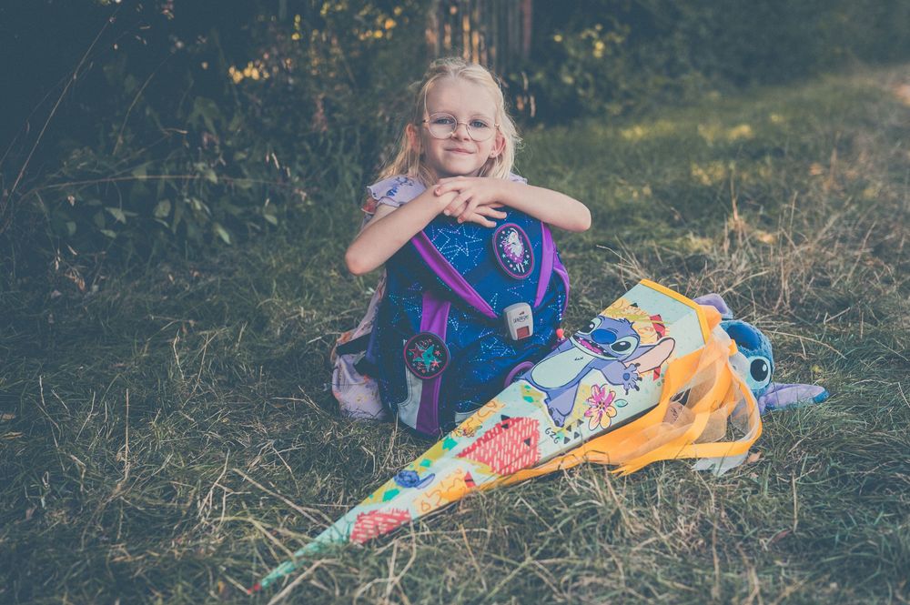 Young blonde girl sitting on hay with colorful school cone and backpack, smiling outdoors in natural setting.