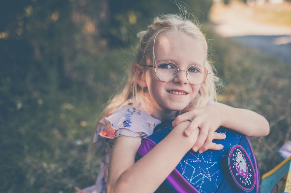 Smiling blonde girl with glasses hugging a colorful school backpack outdoors in a sunny park setting.