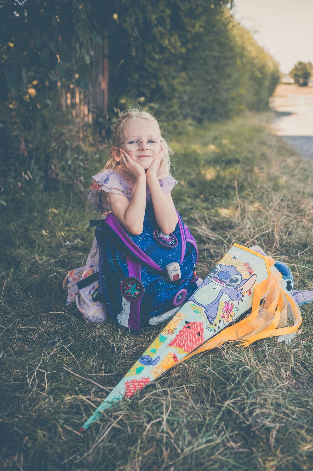 Young blonde girl sitting on grass with colorful school bag and cone-shaped gift bags outdoors.