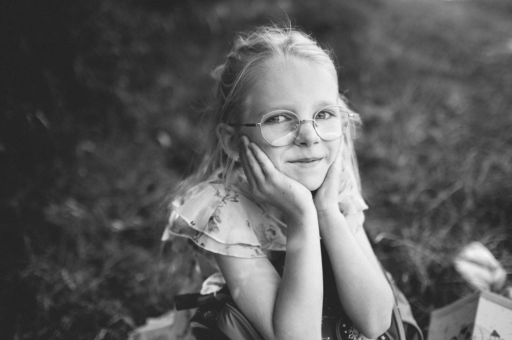 Black and white portrait of a smiling young blonde girl wearing round glasses, resting her chin on her hands outdoors.