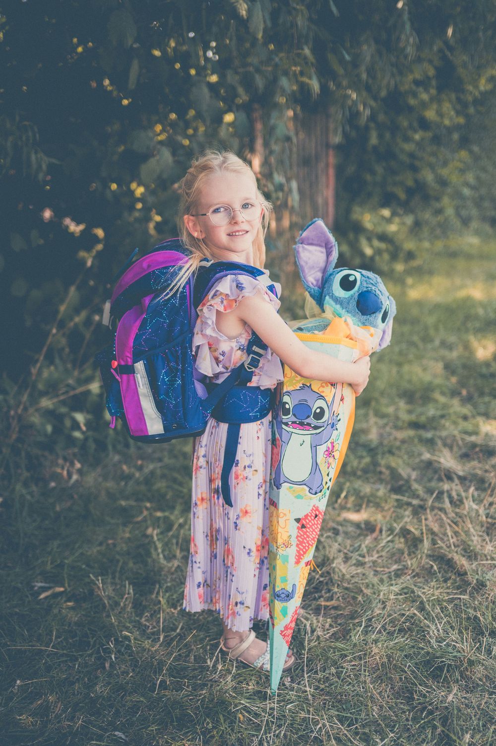 Young girl with blonde hair holding a Stitch stuffed animal and wearing a blue glitter backpack outdoors.