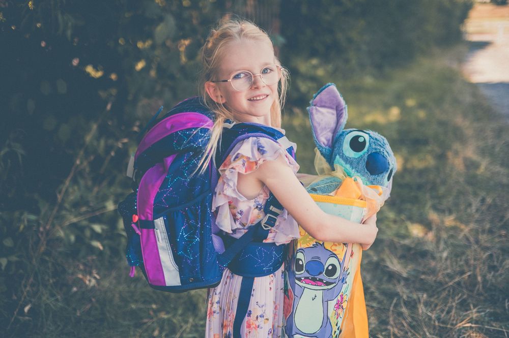 Young blonde girl with glasses holding Stitch plush toy, wearing pink dress and blue backpack outdoors.