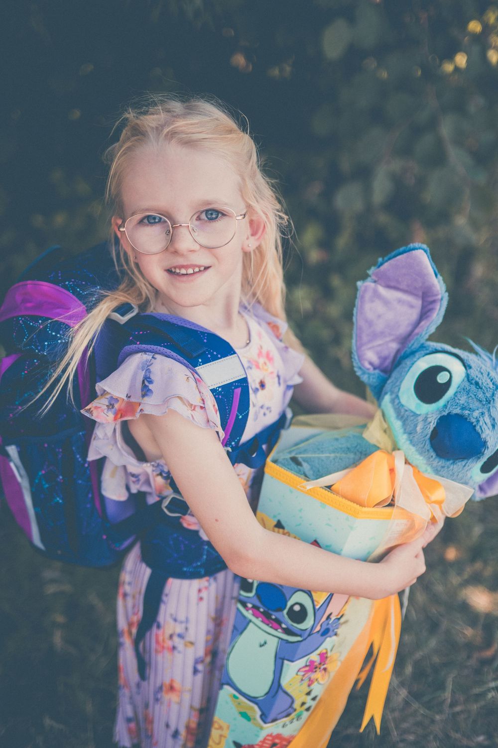 Smiling blonde girl with glasses holding a Stitch plush toy, wearing a floral dress and purple backpack outdoors.