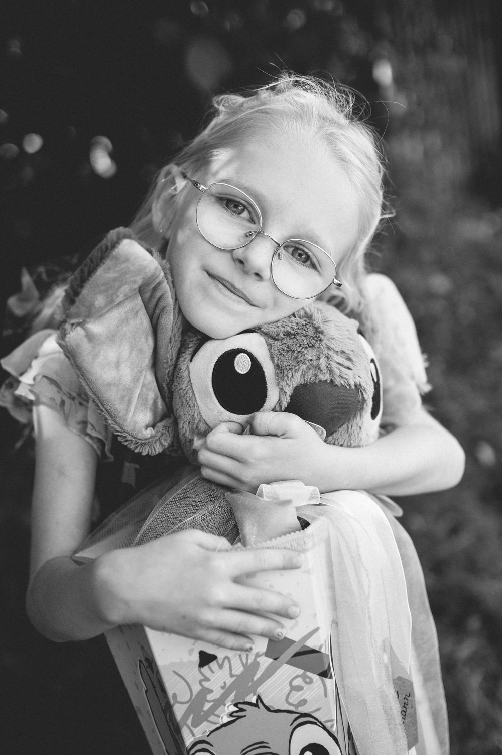 Black and white photo of a smiling young blonde girl with glasses hugging a stuffed animal toy outdoors.