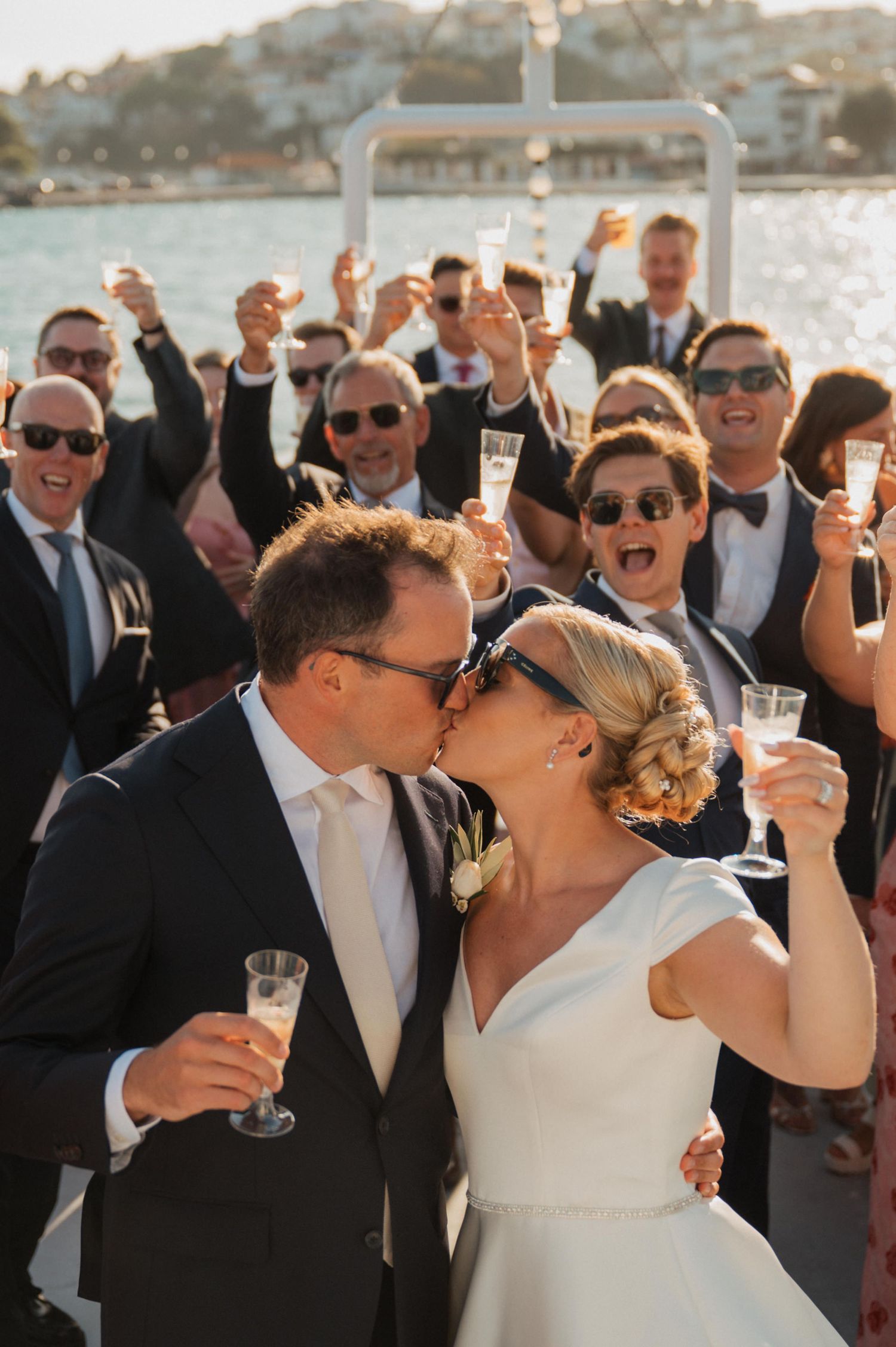 Newlyweds kiss and toast with champagne as wedding guests celebrate on a sunny waterfront venue.