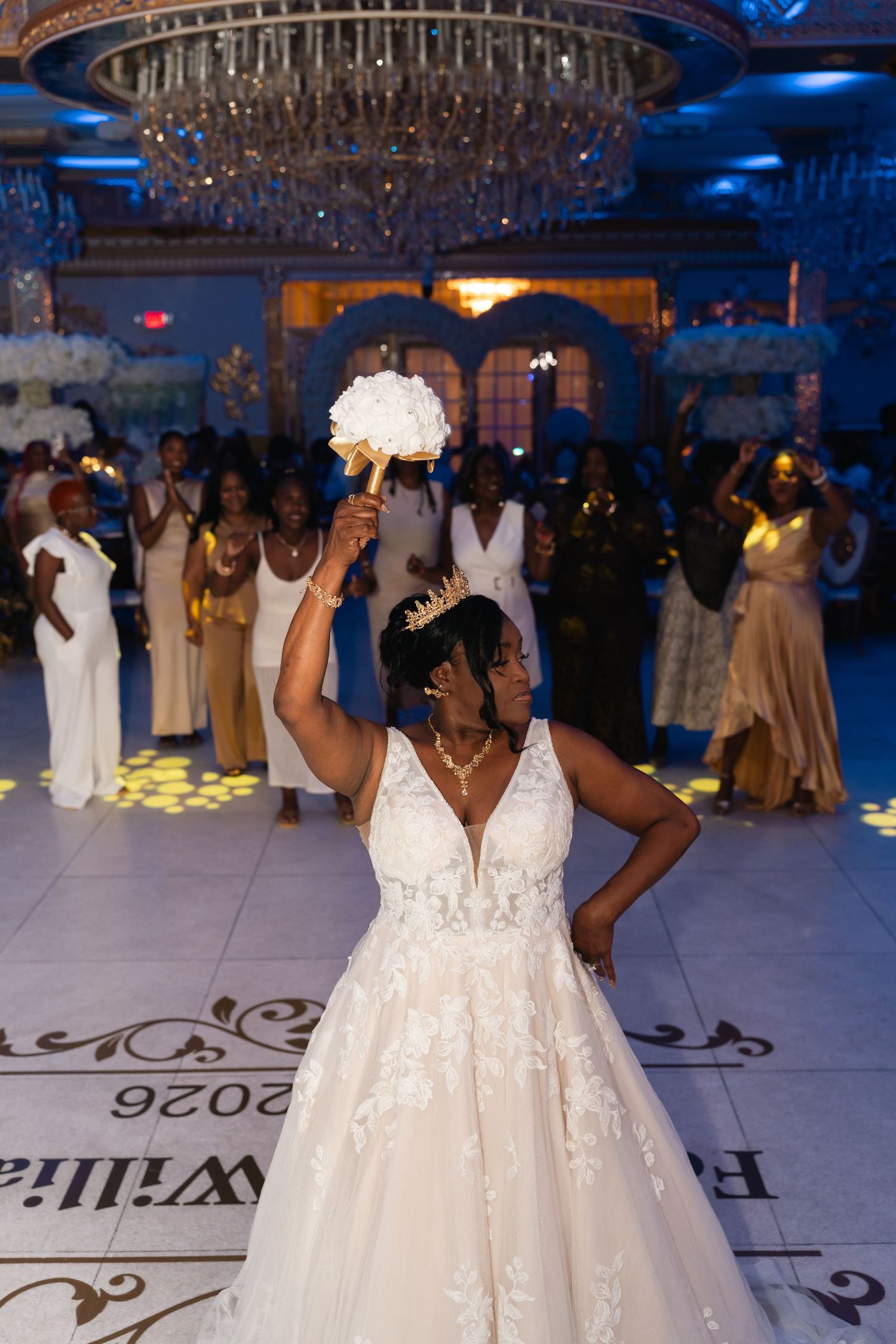 Bride in lace wedding gown tosses bouquet on dance floor at elegant reception with chandelier and blue lighting.