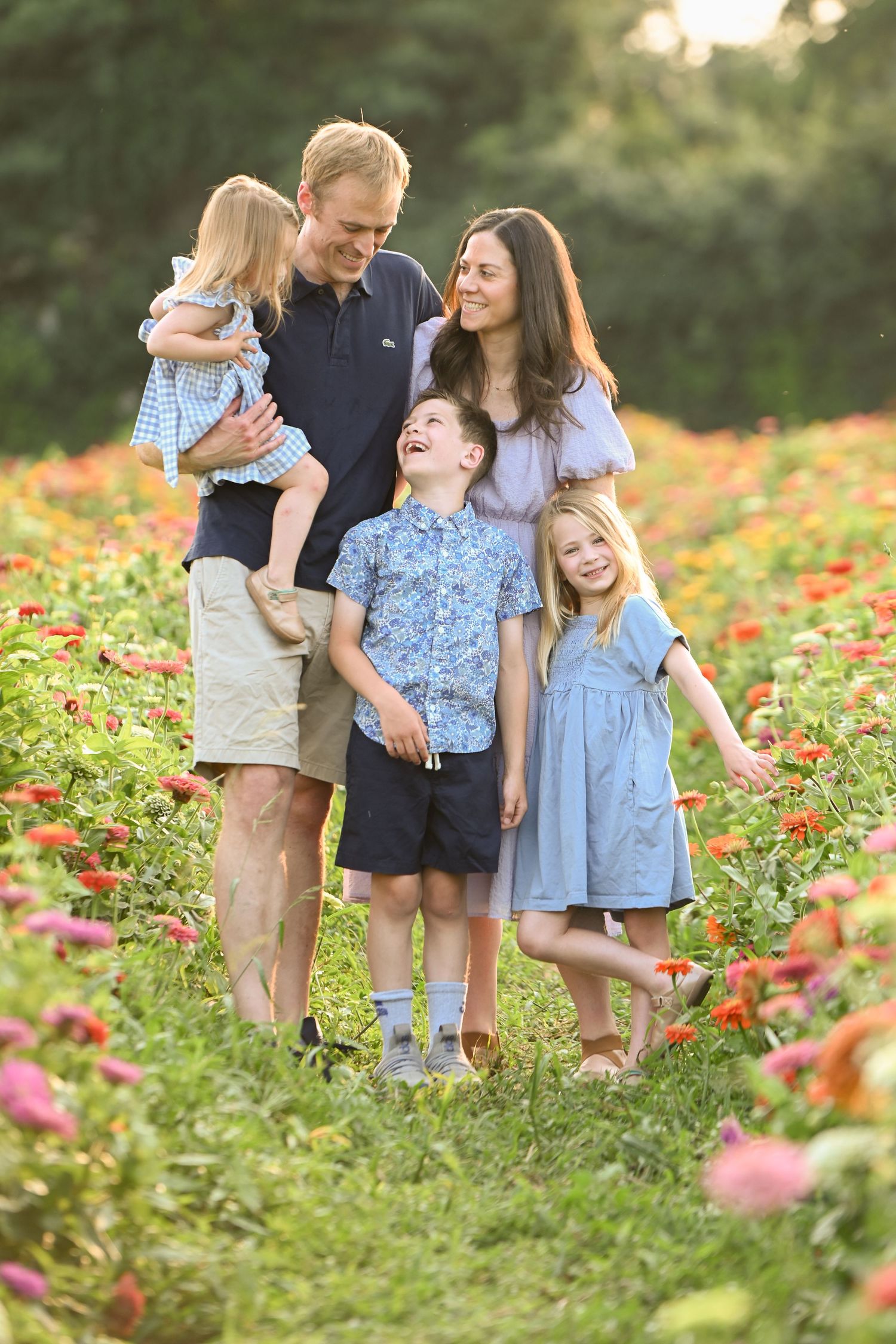 Happy family of five posing in a colorful flower field at golden hour, parents with three young children smiling outdoors.