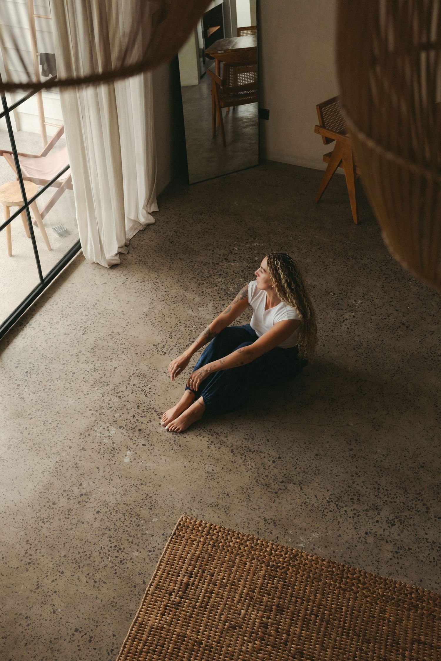 Woman with curly hair sitting barefoot on concrete floor in modern room with sheer curtains and woven rug.