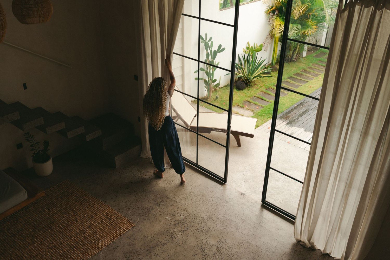 Woman opening large glass French doors leading to a lush tropical garden patio from a modern interior.