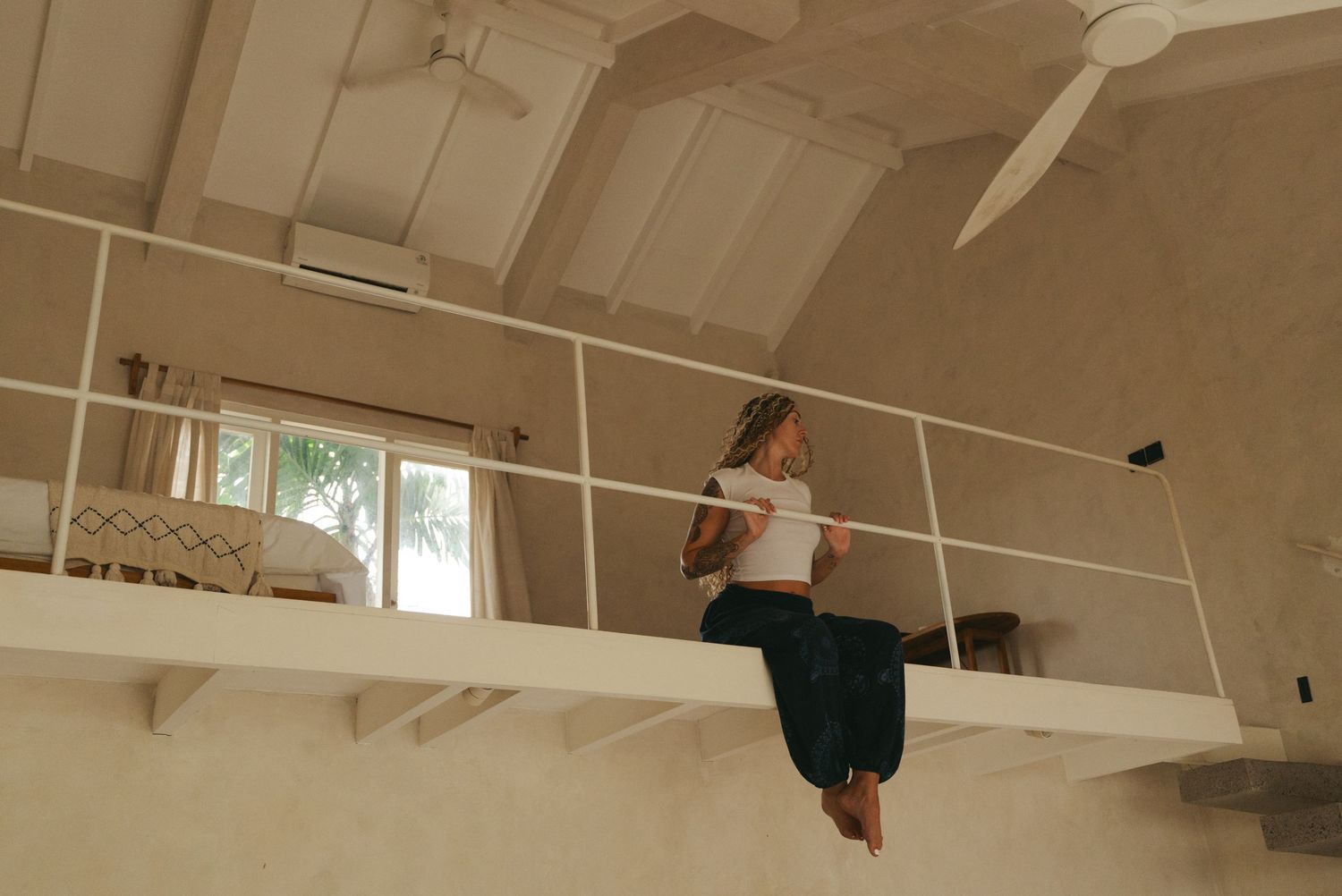 Person sitting on edge of mezzanine loft railing in a bright white minimalist room with vaulted ceiling and ceiling fan.