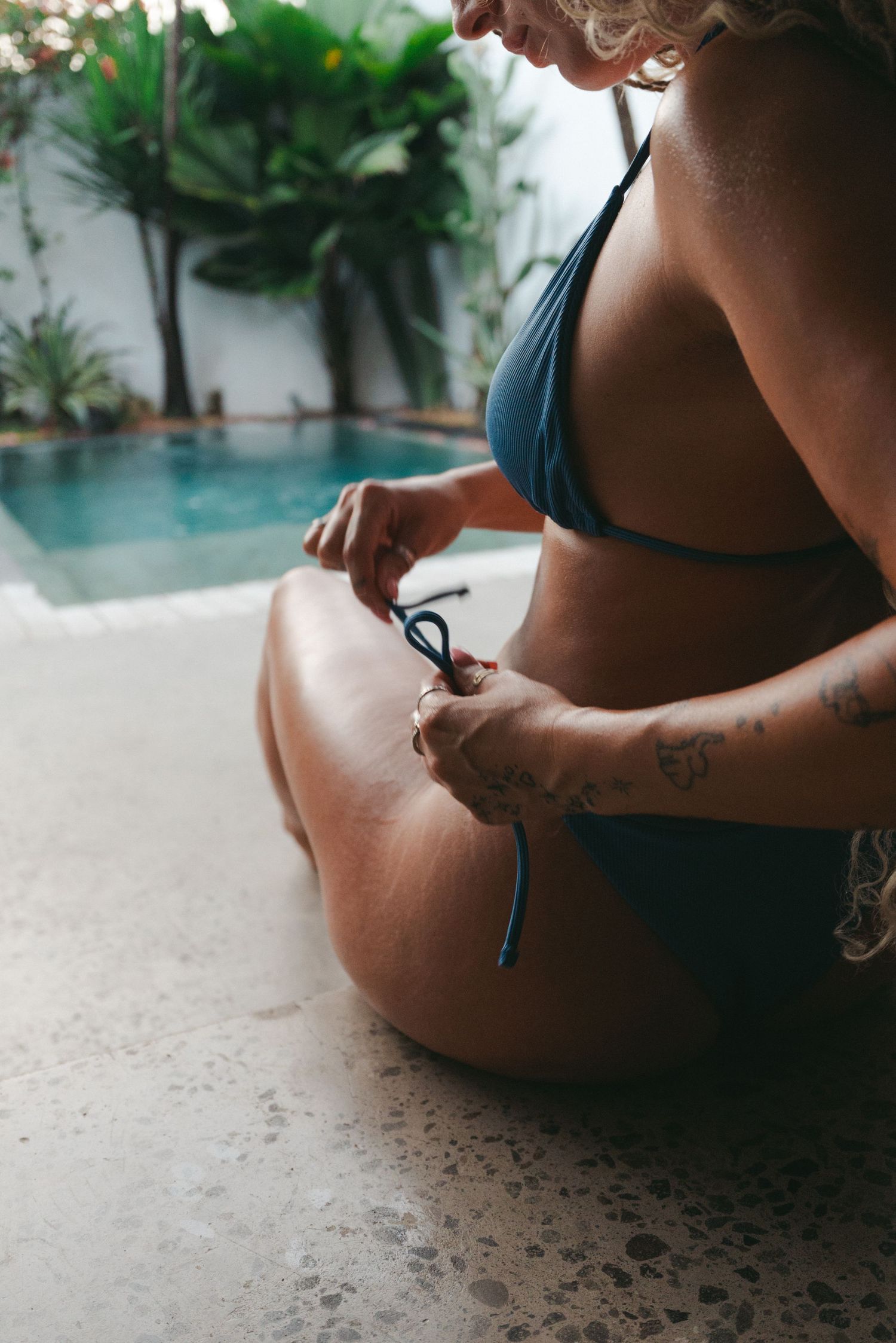 Woman in blue bikini sitting poolside adjusting her swimsuit tie, with tropical plants and pool in background.