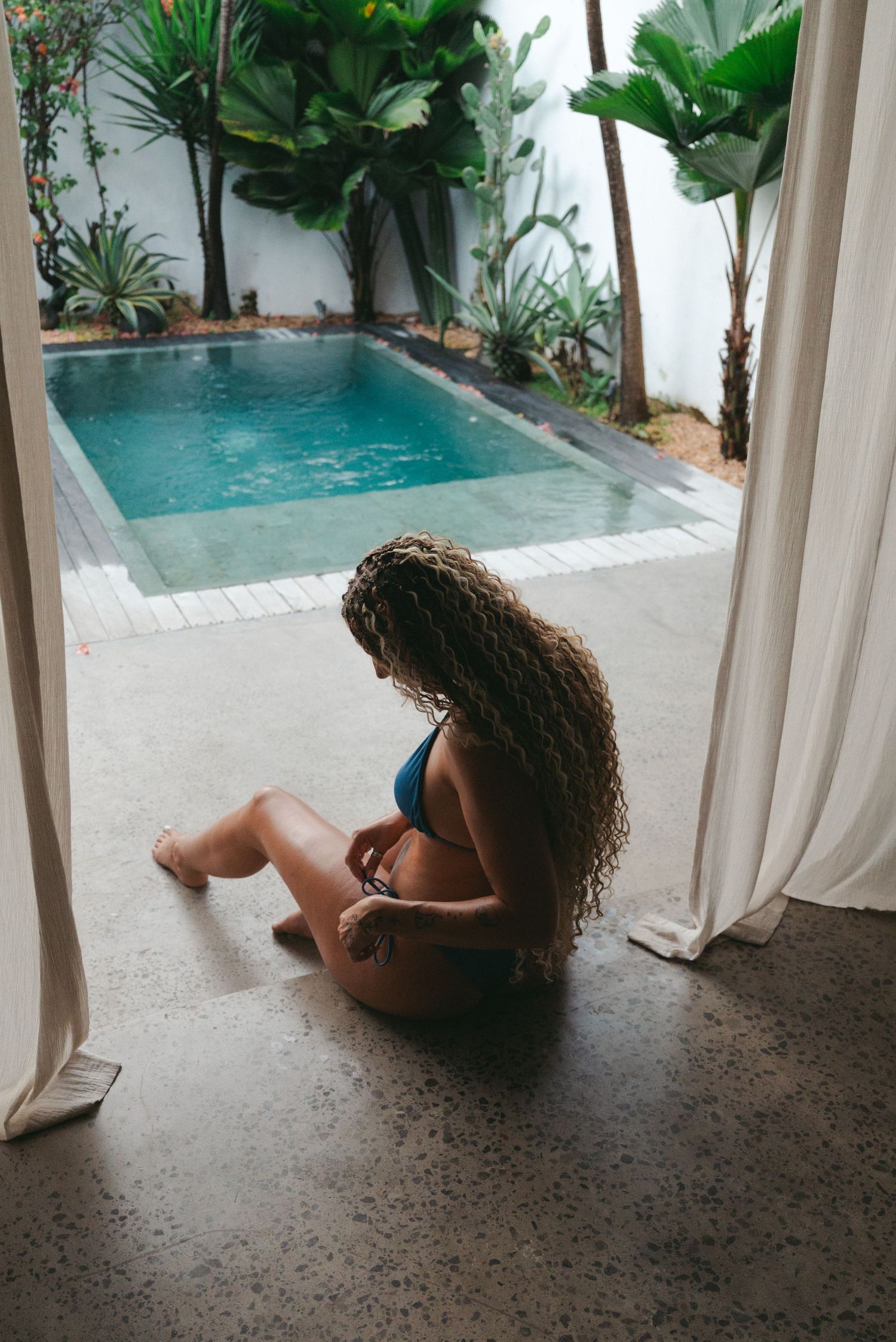 Woman in teal bikini sitting on concrete floor by tropical pool surrounded by lush green plants and white curtains.