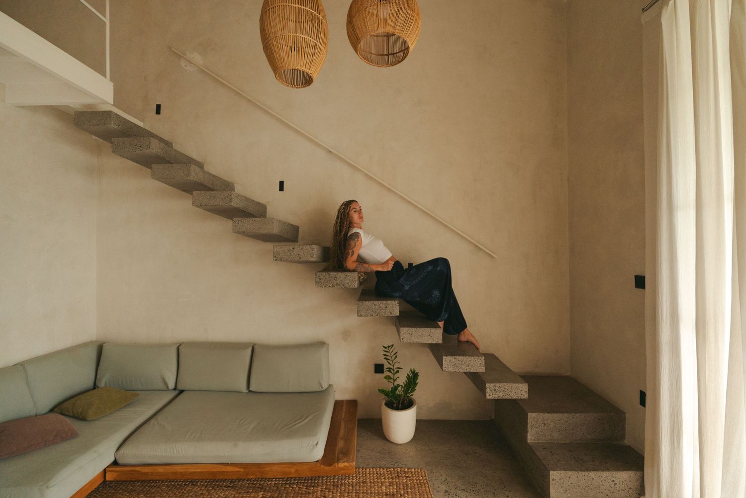 Woman sitting on modern concrete stairs in a minimalist living room with a green sofa and woven pendant lights.