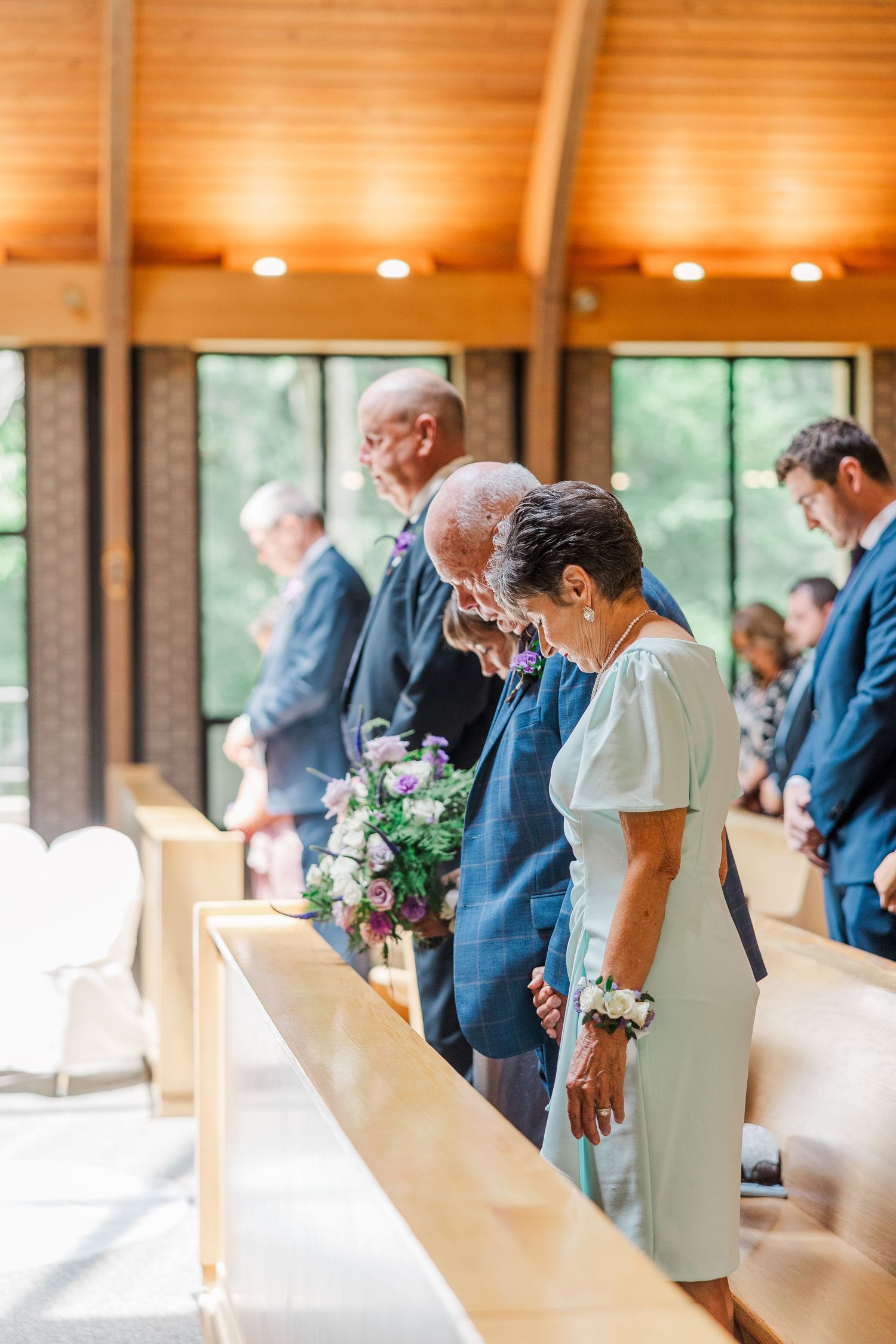 Wedding guests bow heads in prayer during a chapel ceremony, with floral arrangements and wooden pews visible.
