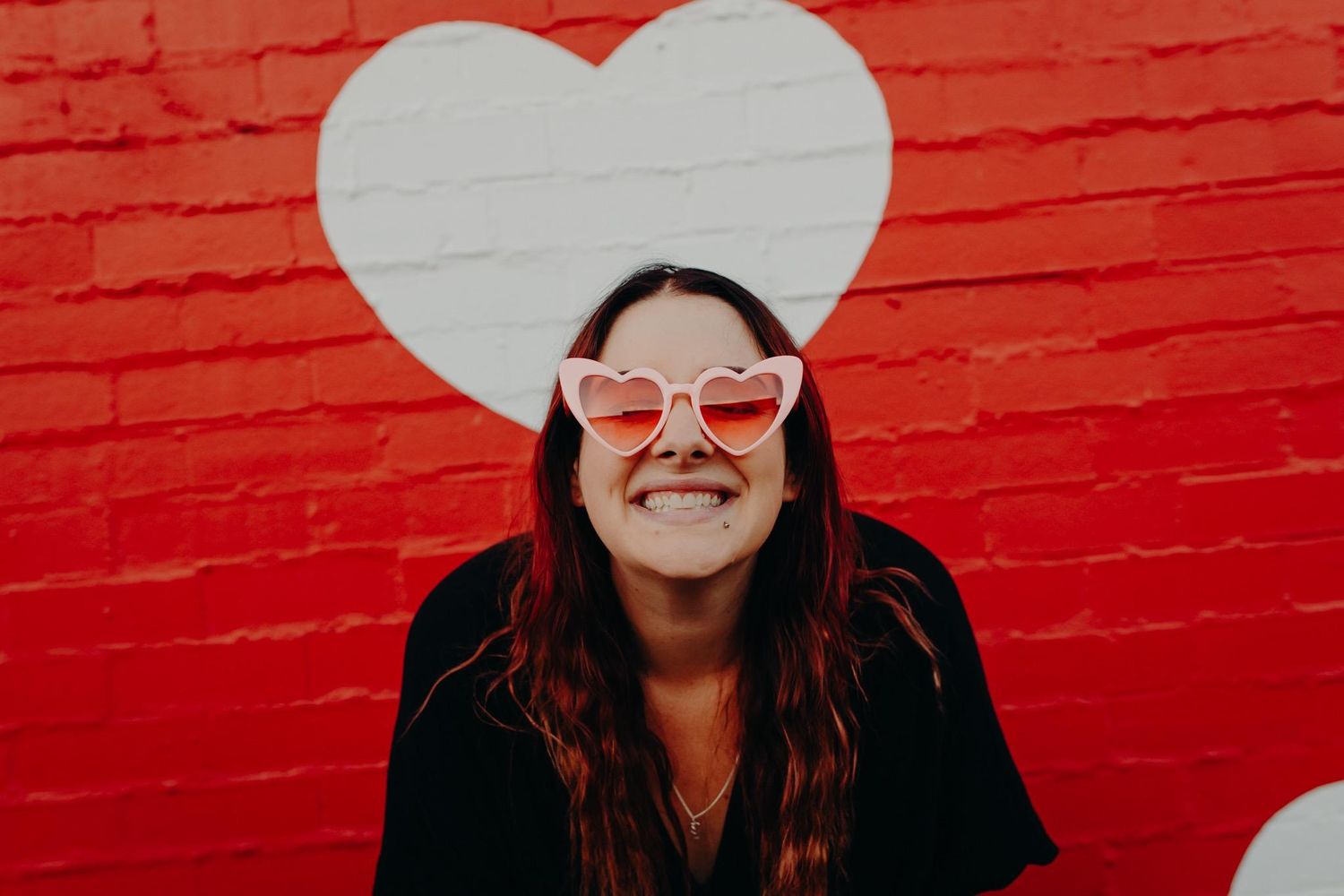 Person in sunglasses poses against red wall with white heart shapes.