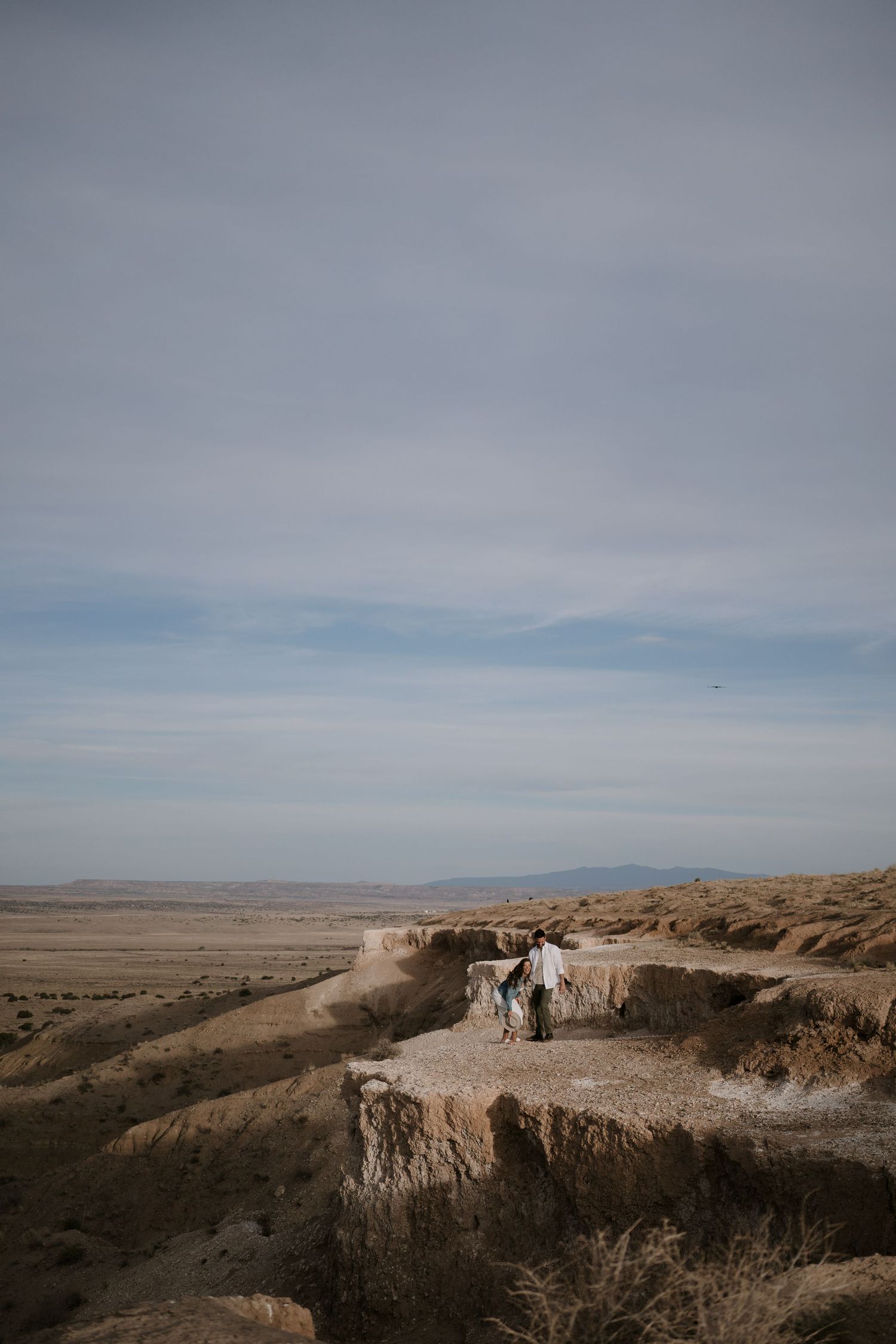 Two people stand on a rocky desert cliff edge overlooking a vast arid landscape under a cloudy sky.