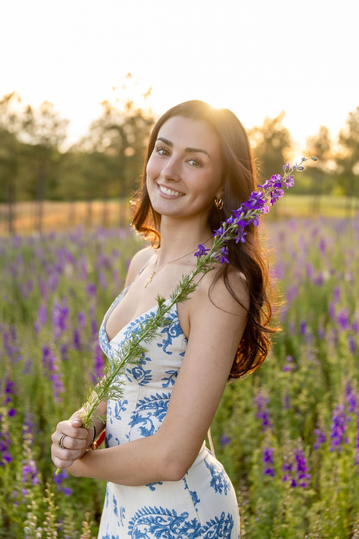 Young woman in blue floral dress holding purple wildflowers, smiling in a lavender field at golden hour sunset.