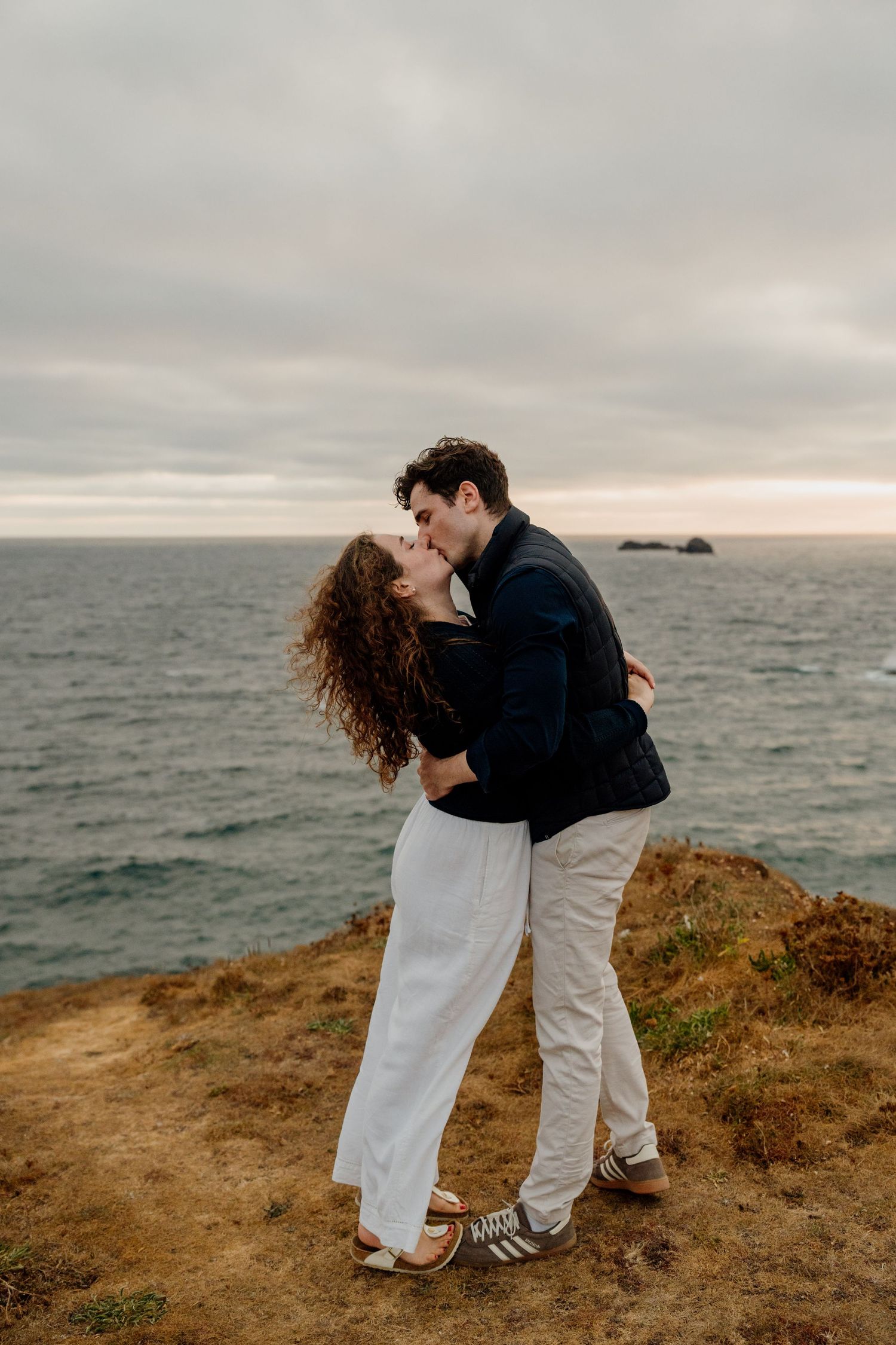 Couple kissing on coastal cliff with ocean backdrop, woman in white outfit, man in navy jacket, romantic engagement photo.