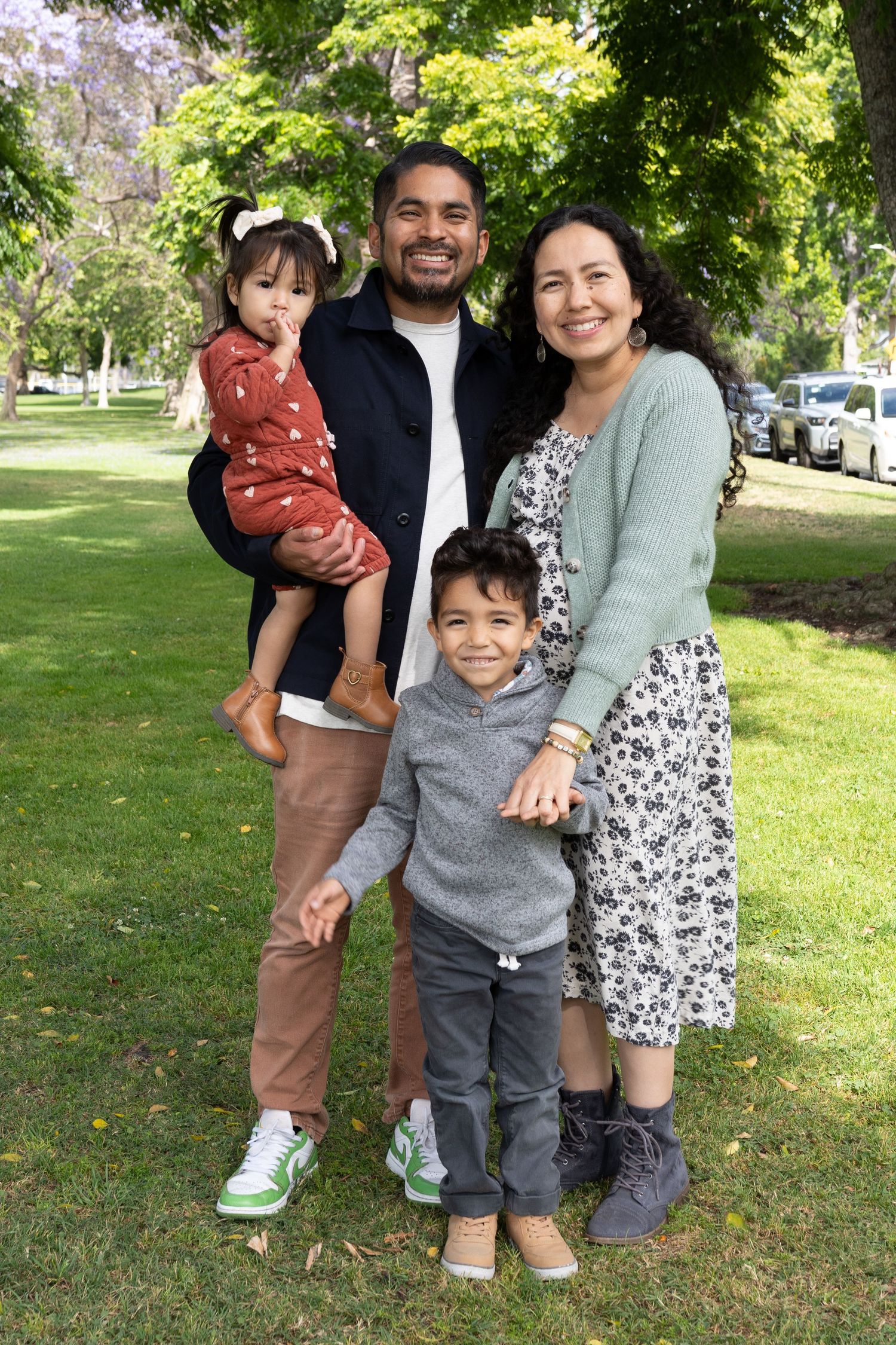 Happy family of four posing outdoors in a green park, parents with two young children smiling on a sunny day.