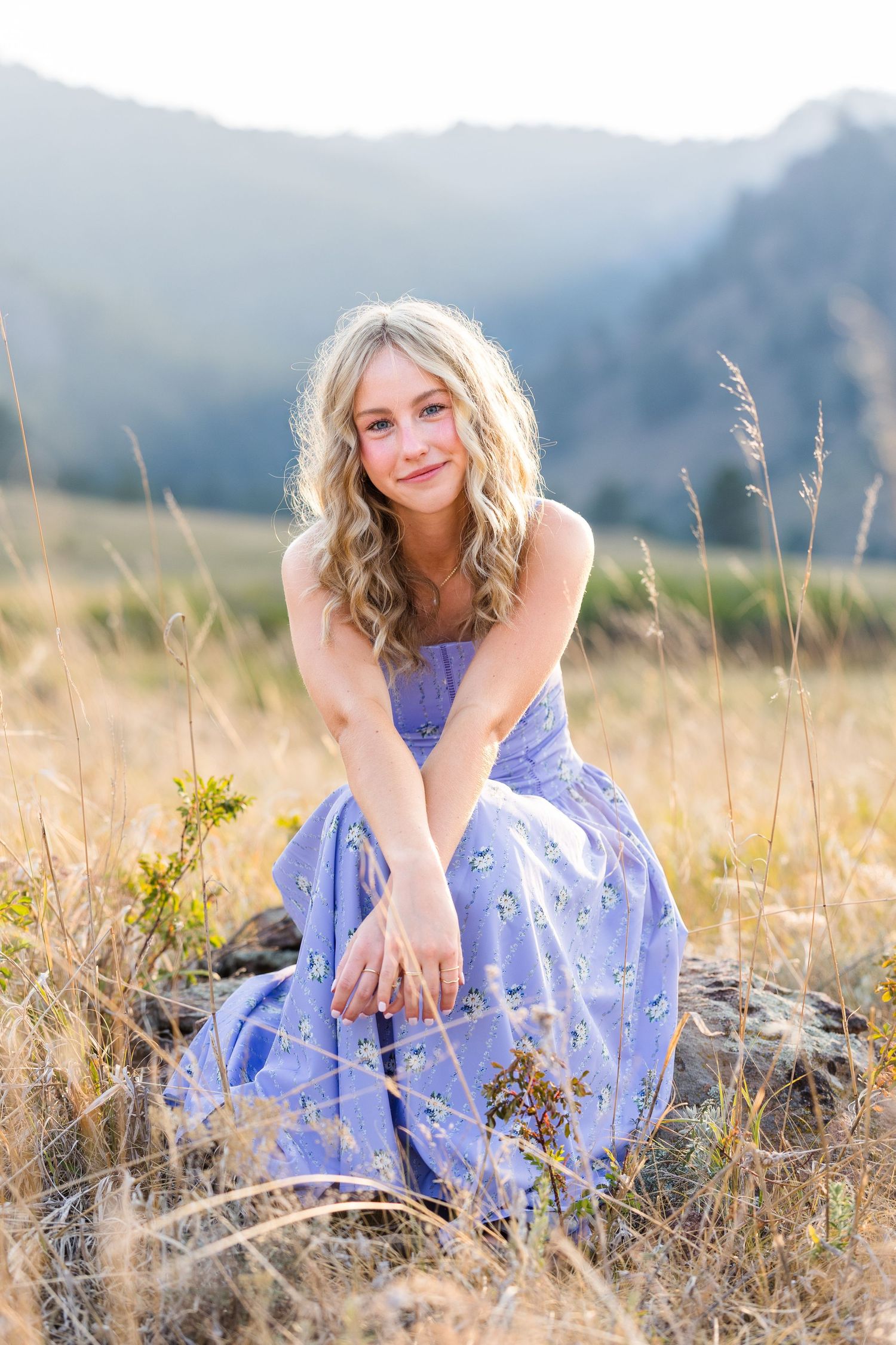 Young blonde woman in lavender floral dress sitting in golden meadow with mountain backdrop in warm sunlight.