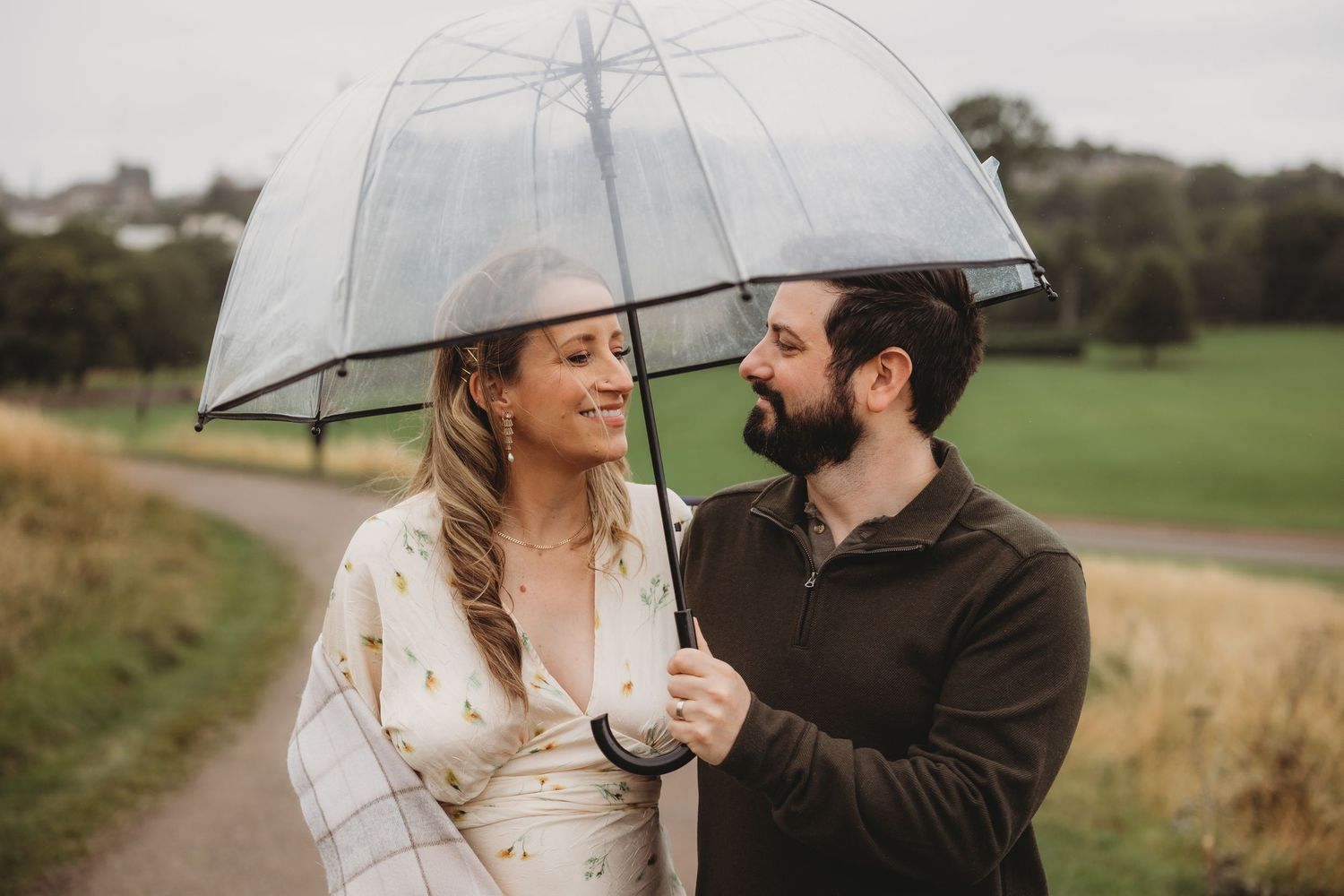 Couple sharing a clear umbrella in the rain, smiling at each other in a lush green park setting.