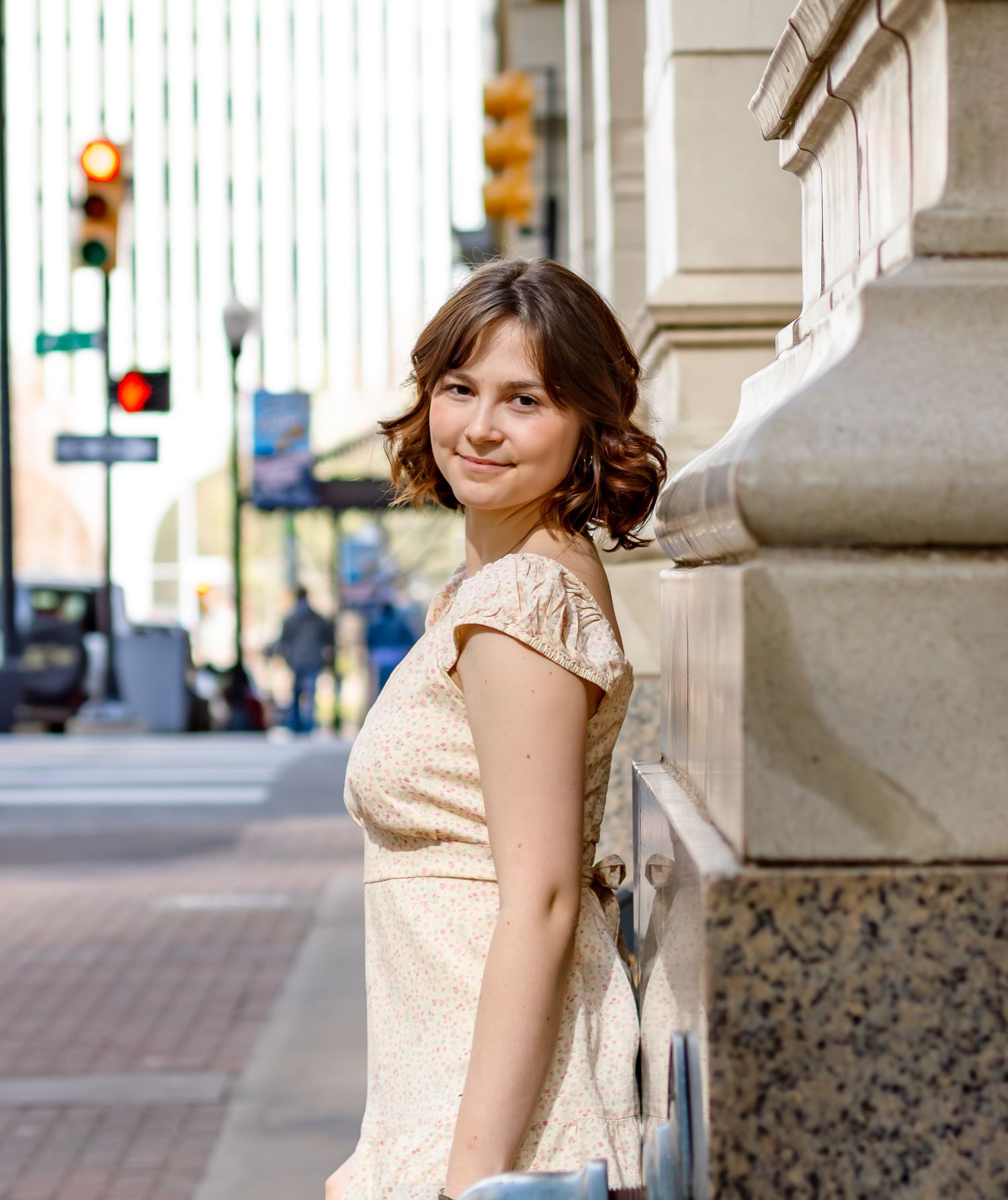 Young woman in a cream lace dress smiling over her shoulder on a sunny urban sidewalk near a stone building.