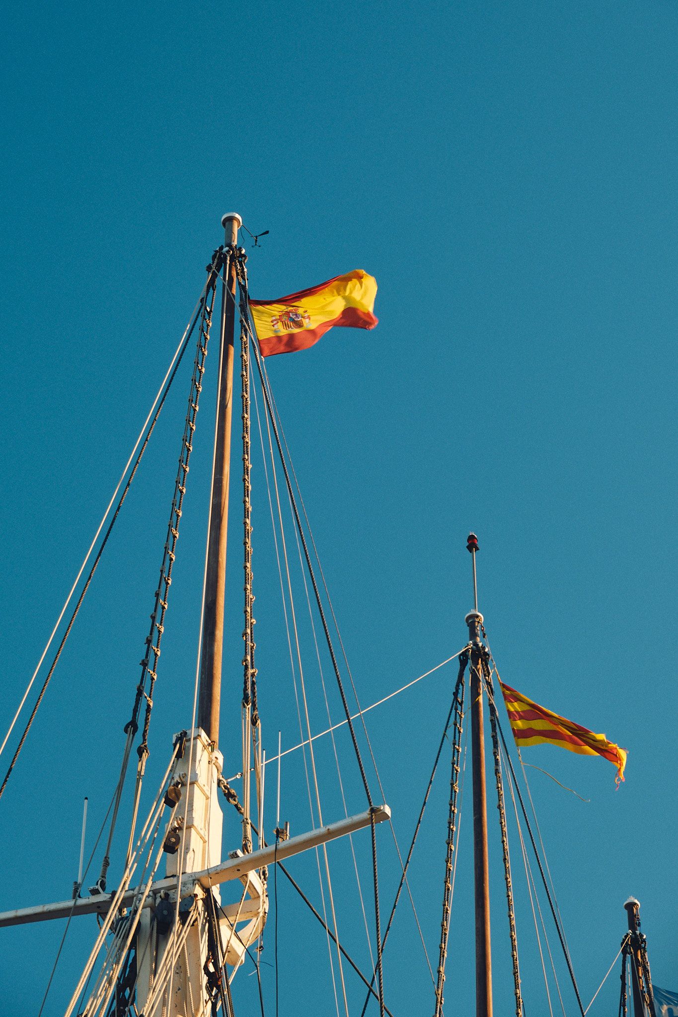 Spanish and Catalan flags flying on tall sailing ship masts against a clear blue sky.