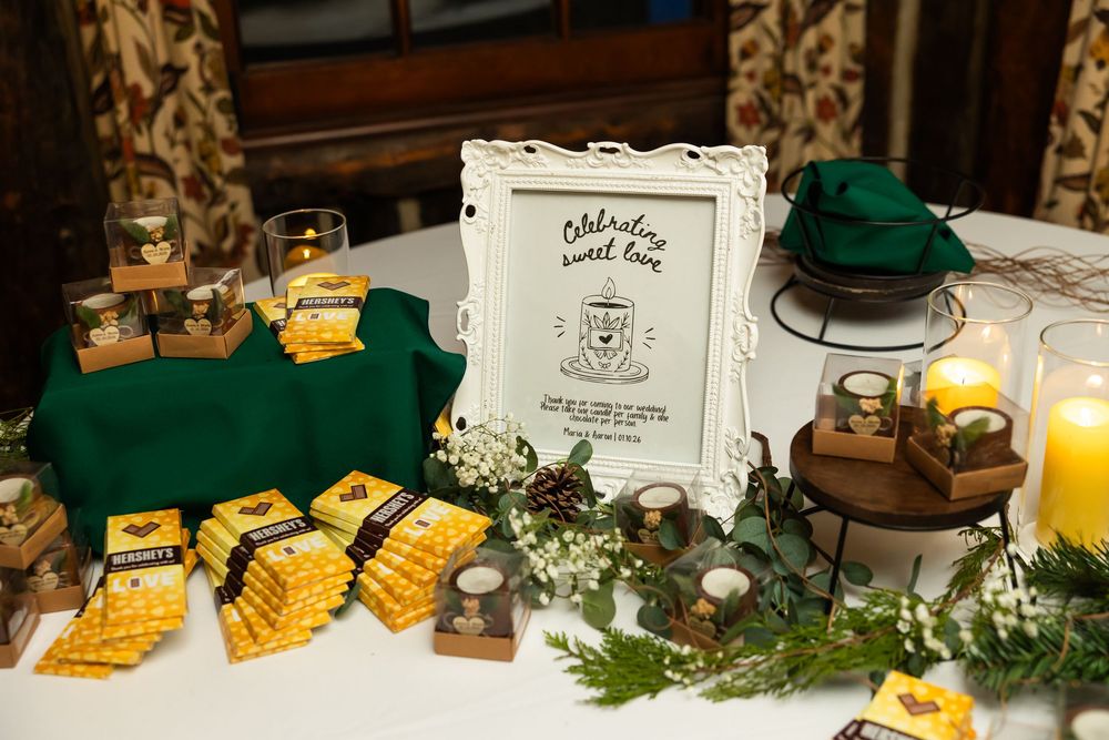 Elegant wedding dessert table with green linens, yellow packaged treats, candles, and a decorative framed sign celebrating sweet love.