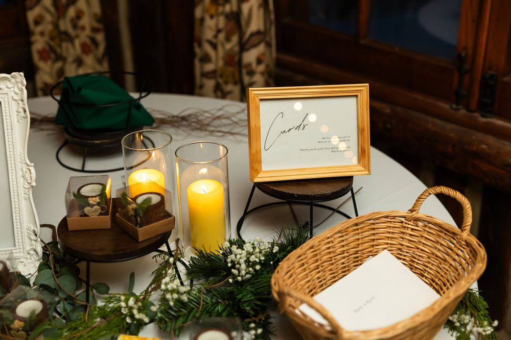 Wedding card table with lit candles, framed cards sign, wicker basket, and floral greenery decorations.