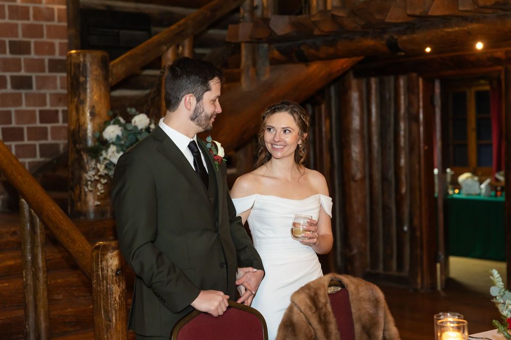 Bride in off-shoulder gown and groom in dark suit smile during rustic log cabin wedding reception.