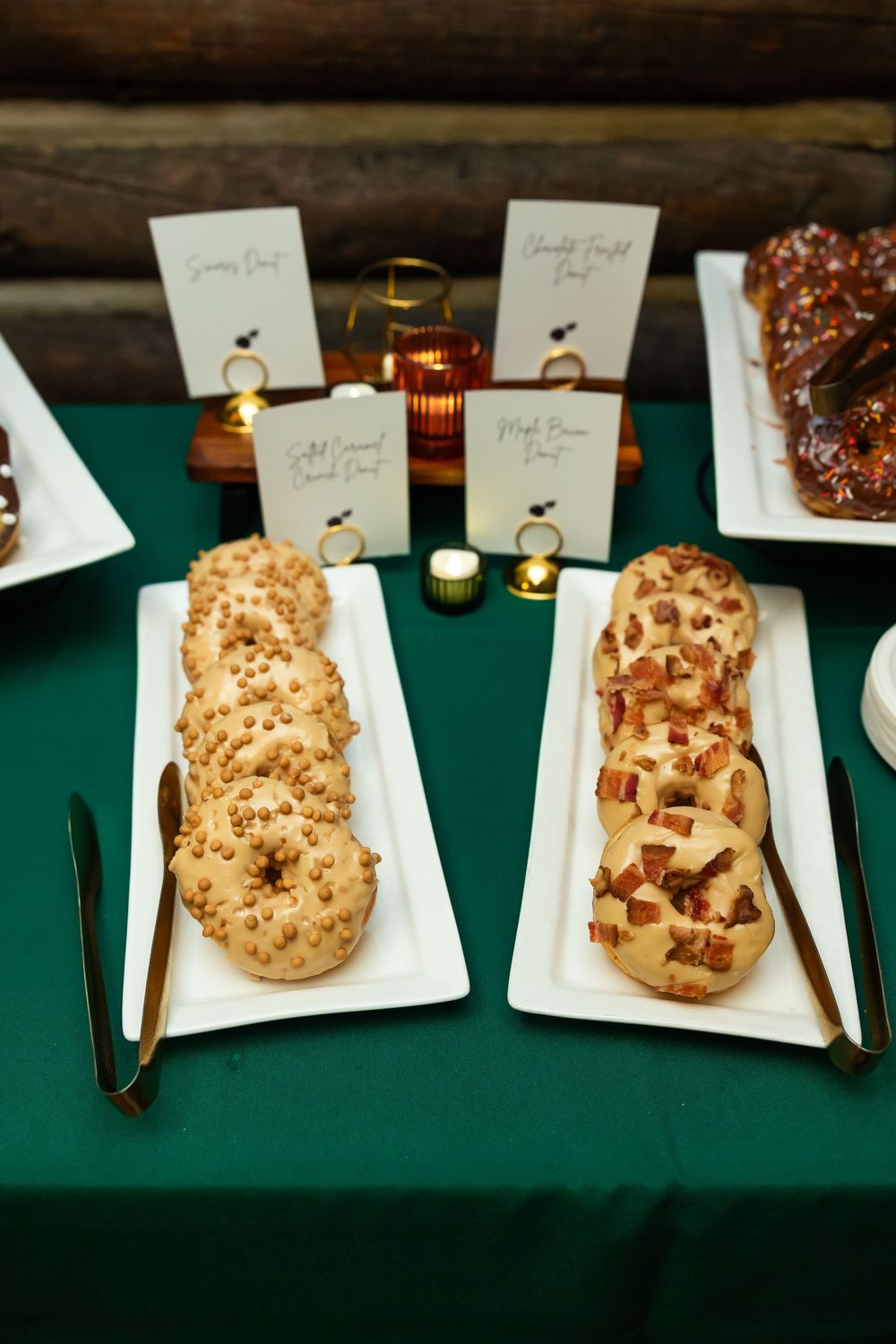 Elegant donut display on green tablecloth with white platters, gold utensils, and decorative name cards at an event.