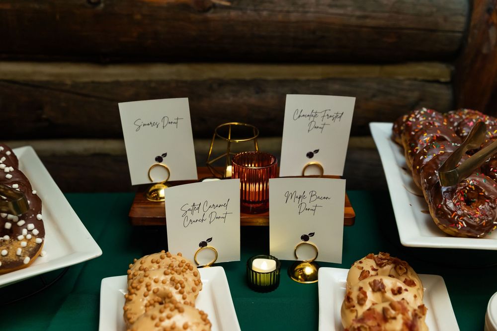 Elegant dessert table display with labeled pastries and donuts on white platters, accented by candlelight and green tablecloth.
