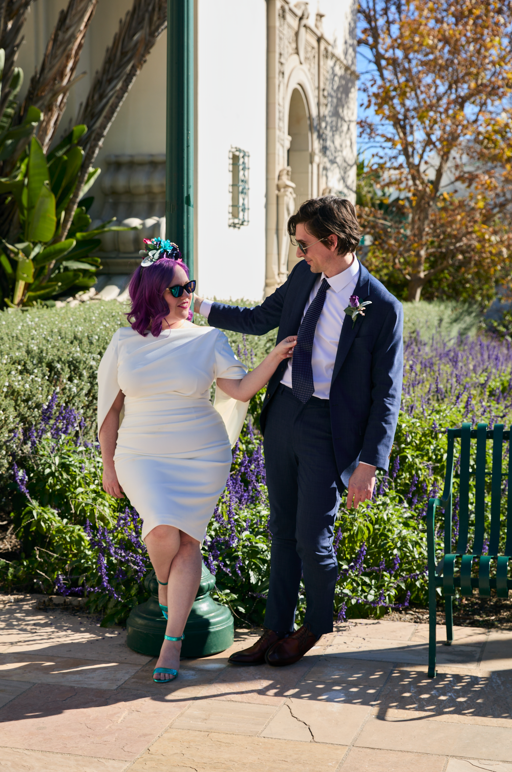 A couple in wedding attire, woman in white dress with purple hair, man in navy suit, posing outdoors among purple flowers.