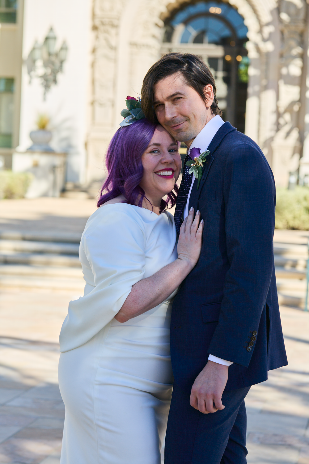 A bride with purple hair in a white dress and groom in navy suit pose together outside a building on their wedding day.