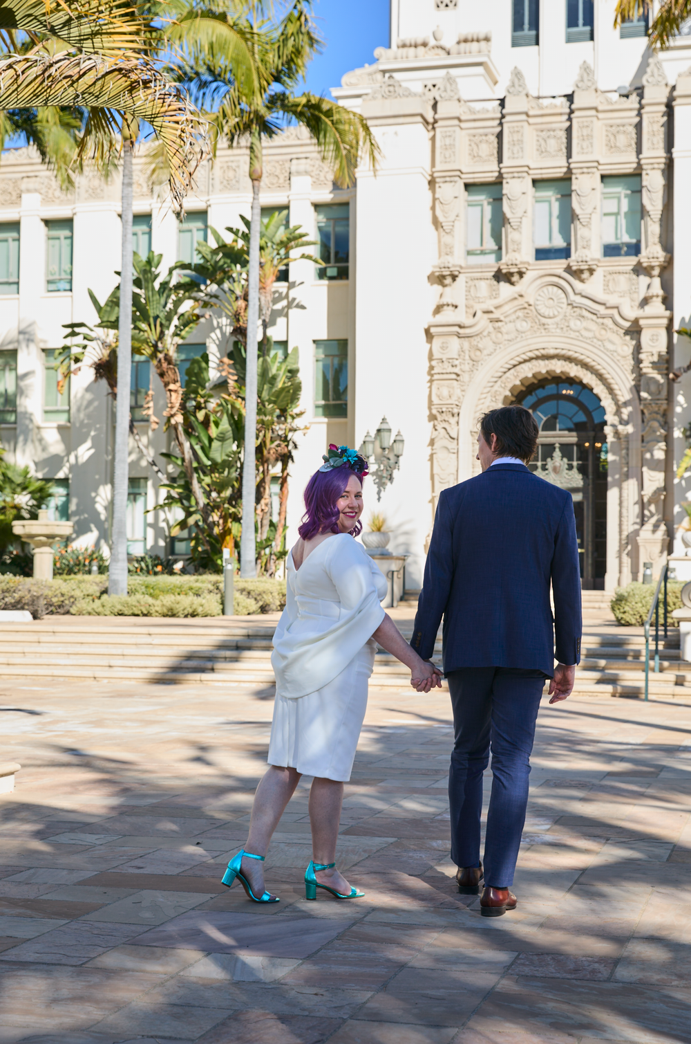 Couple holding hands walking toward ornate white building with palm trees, bride in white dress with purple hair and teal heels.