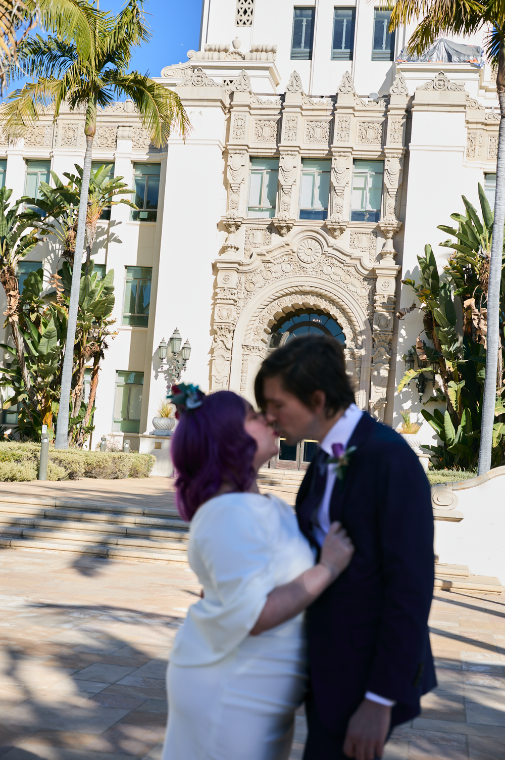 Couple shares a romantic kiss in wedding attire outside an ornate building with palm trees in San Diego.
