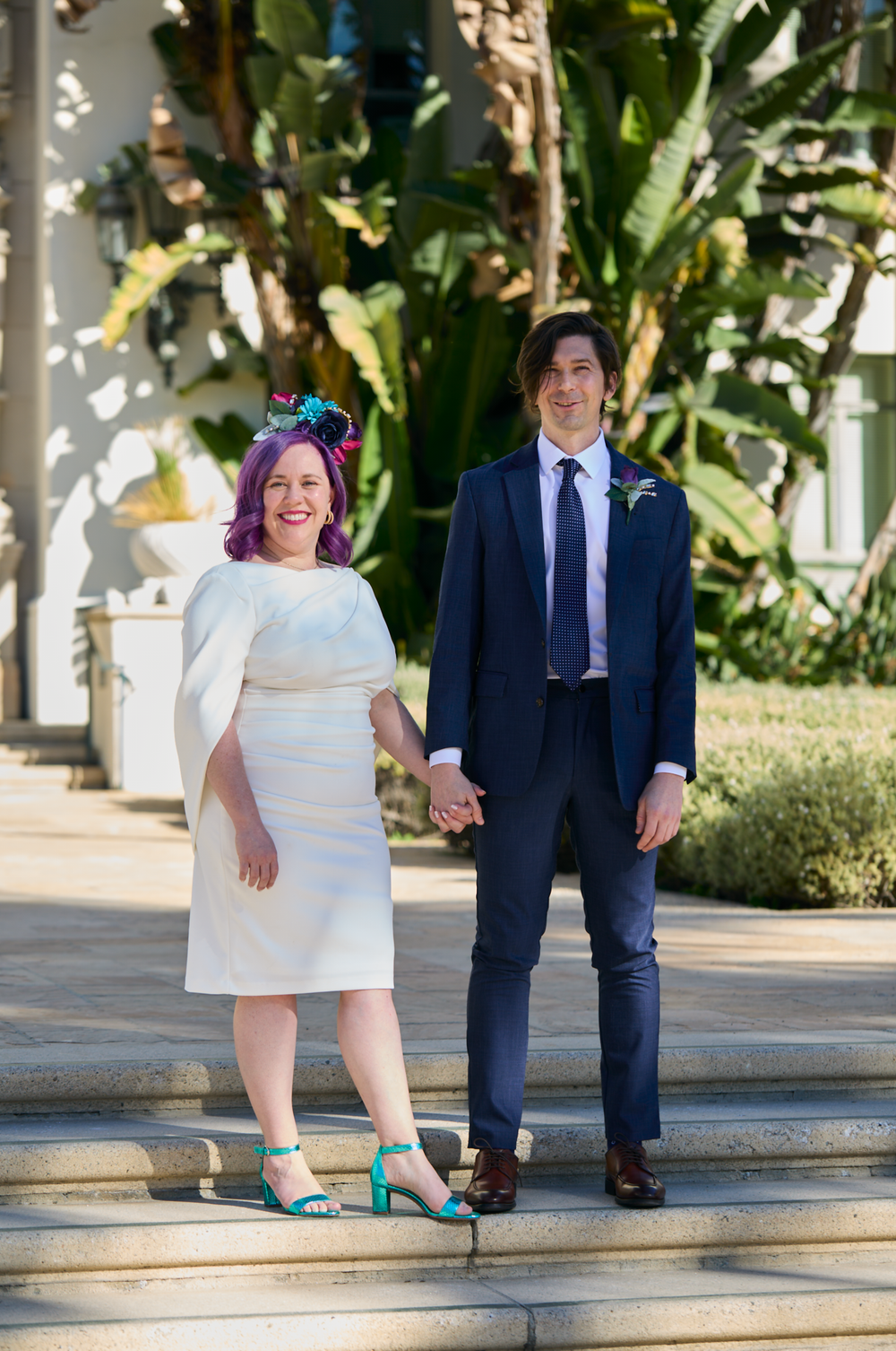 A bride with purple hair and teal heels holds hands with a groom in a navy suit on outdoor steps.