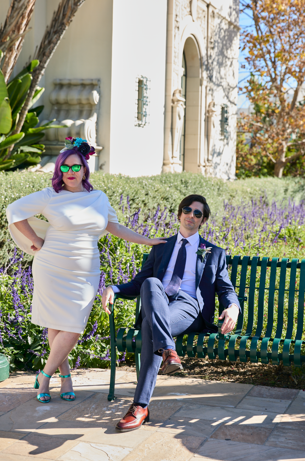 Bride in white dress and groom in navy suit pose by a green bench in a garden setting.