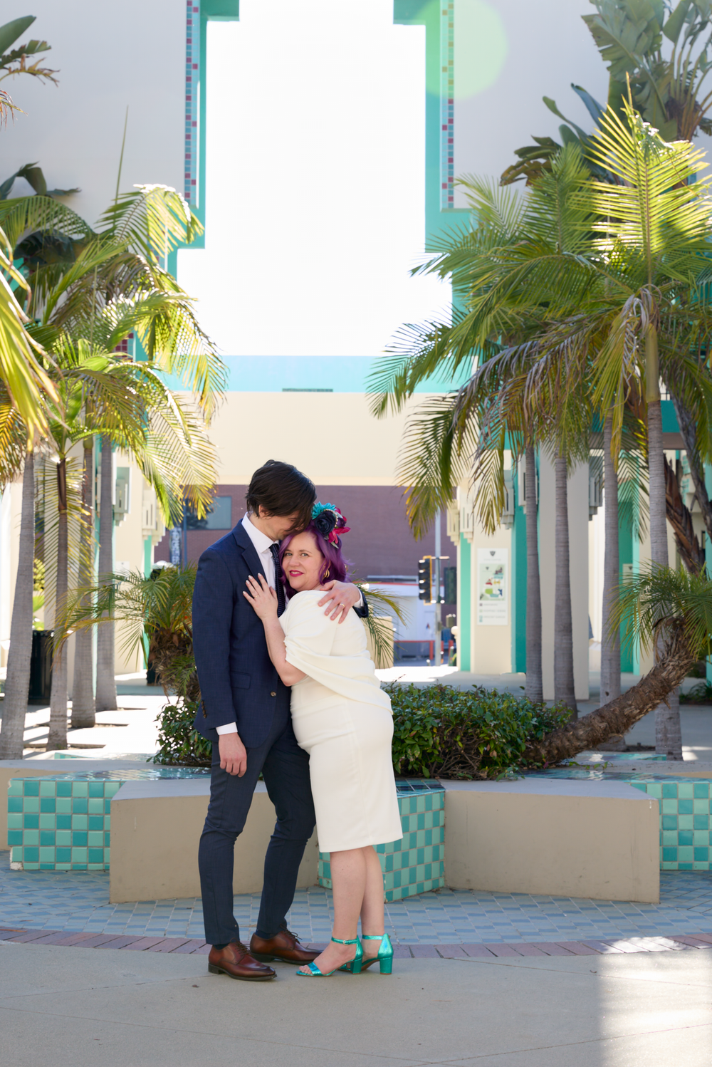 Couple embracing outdoors near palm trees and colorful Art Deco architecture, woman in white dress and teal heels.
